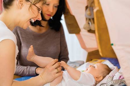 Quelle: © Kzenon_shutterstock Eine Hebamme hält die Beine eines vor ihr liegenden Säuglings und lacht ihn an
