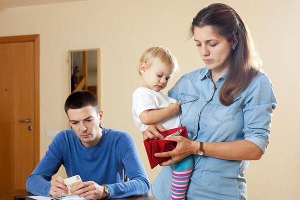Ein Mann sitzt am Tisch, neben ihm steht eine Frau mit Kind auf den Arm und schaut in einen leeren Geldbeutel